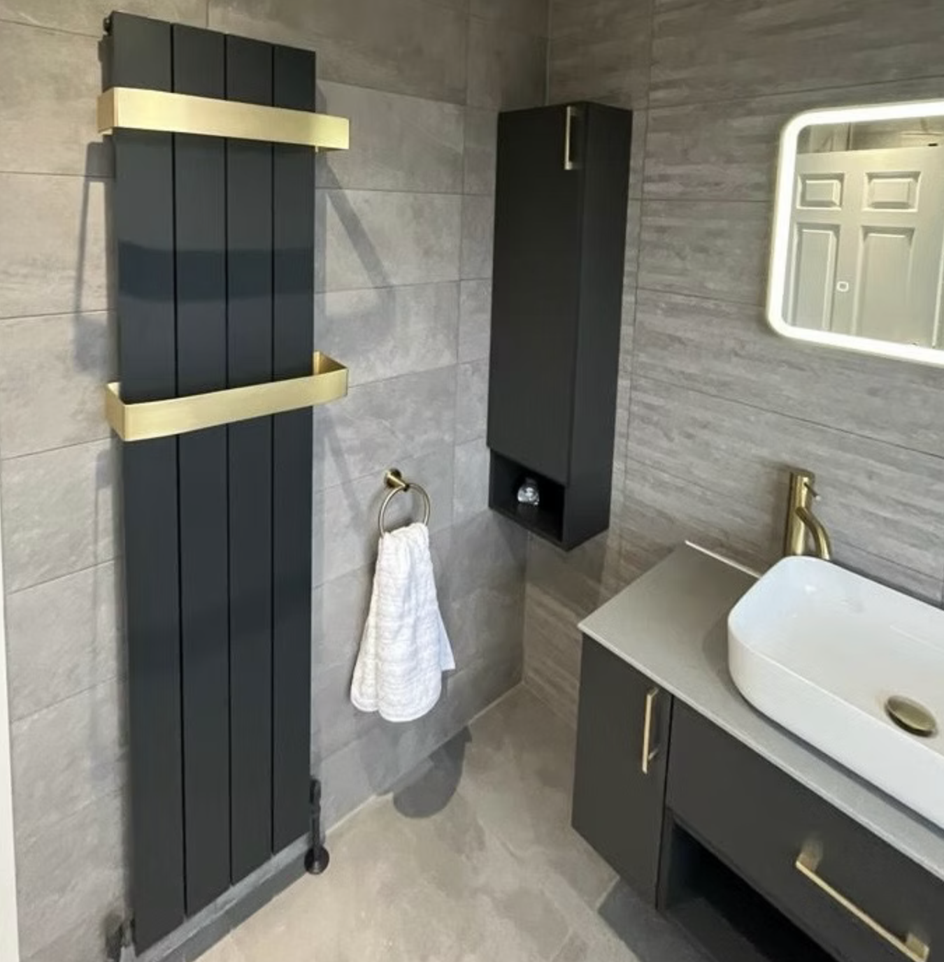 Detail: an anthracite vertical radiator with brushed brass towel rails, brass tap and a charcoal wall-hung vanity against grey stone-effect tiles.