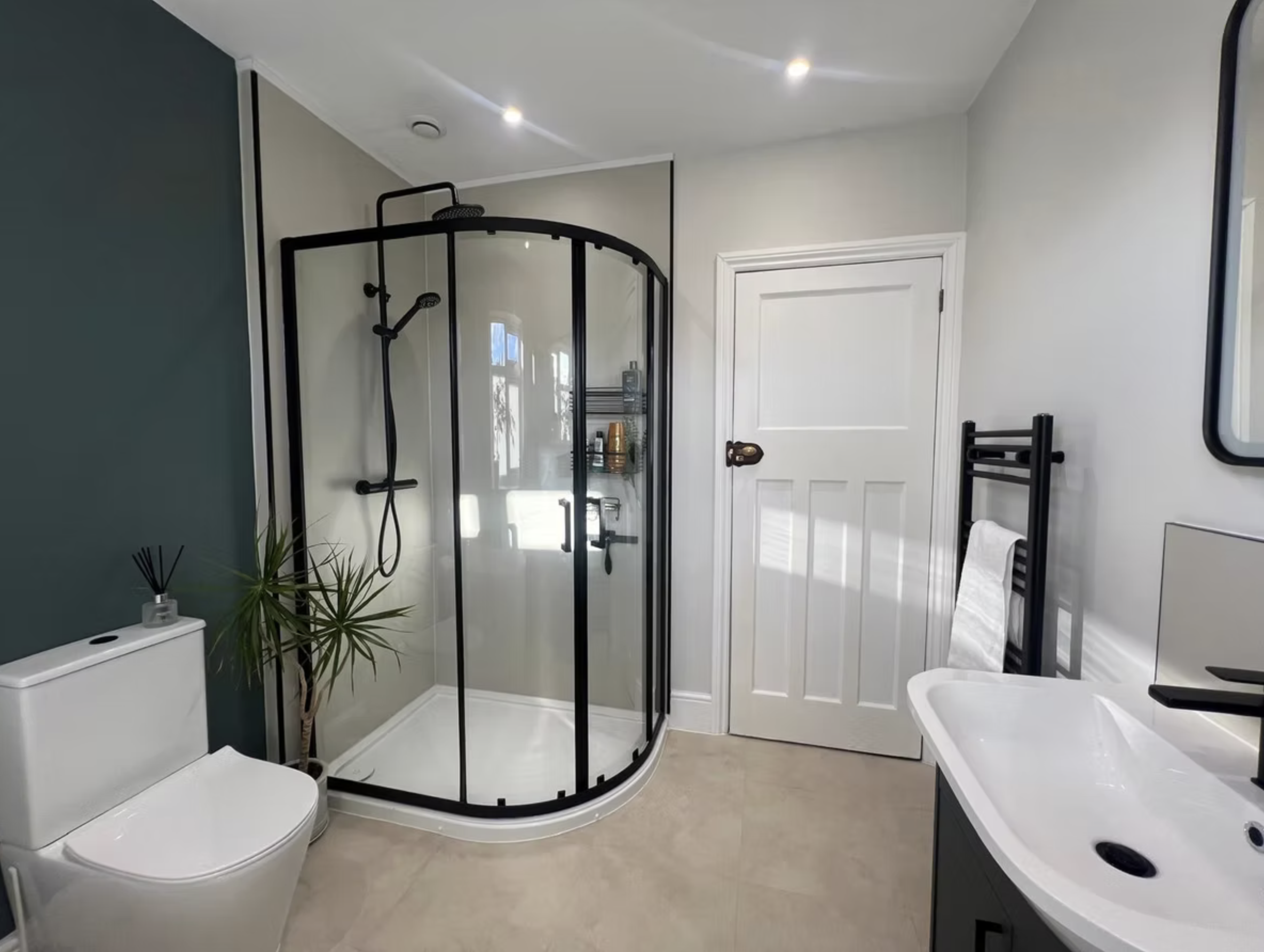 A carefully finished bathroom with a black-framed shower, warm stone flooring and a deep green feature wall.