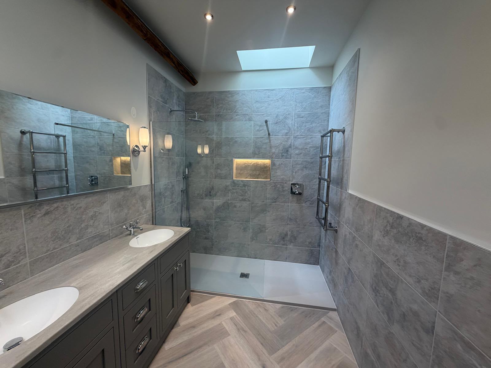 A spacious grey bathroom with a double vanity, frameless walk-in shower, illuminated niche and rooflight.