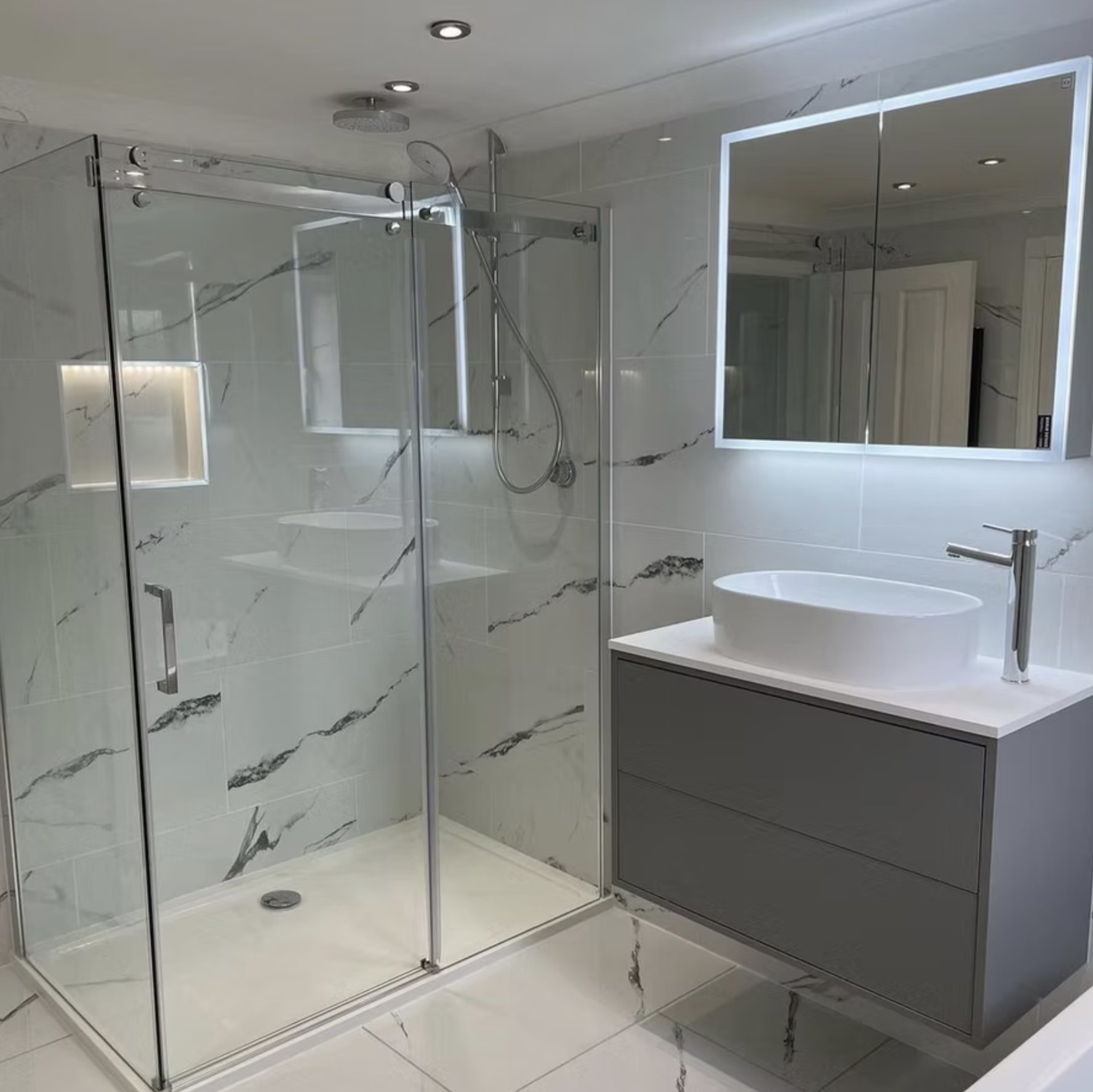 The walk-in shower behind frameless sliding glass, with a lit recess and the grey vanity beyond.