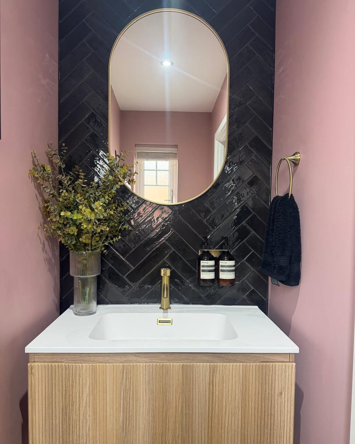 A pink bathroom vanity with glossy black herringbone tiles, an arched brass mirror and fluted oak cabinetry.