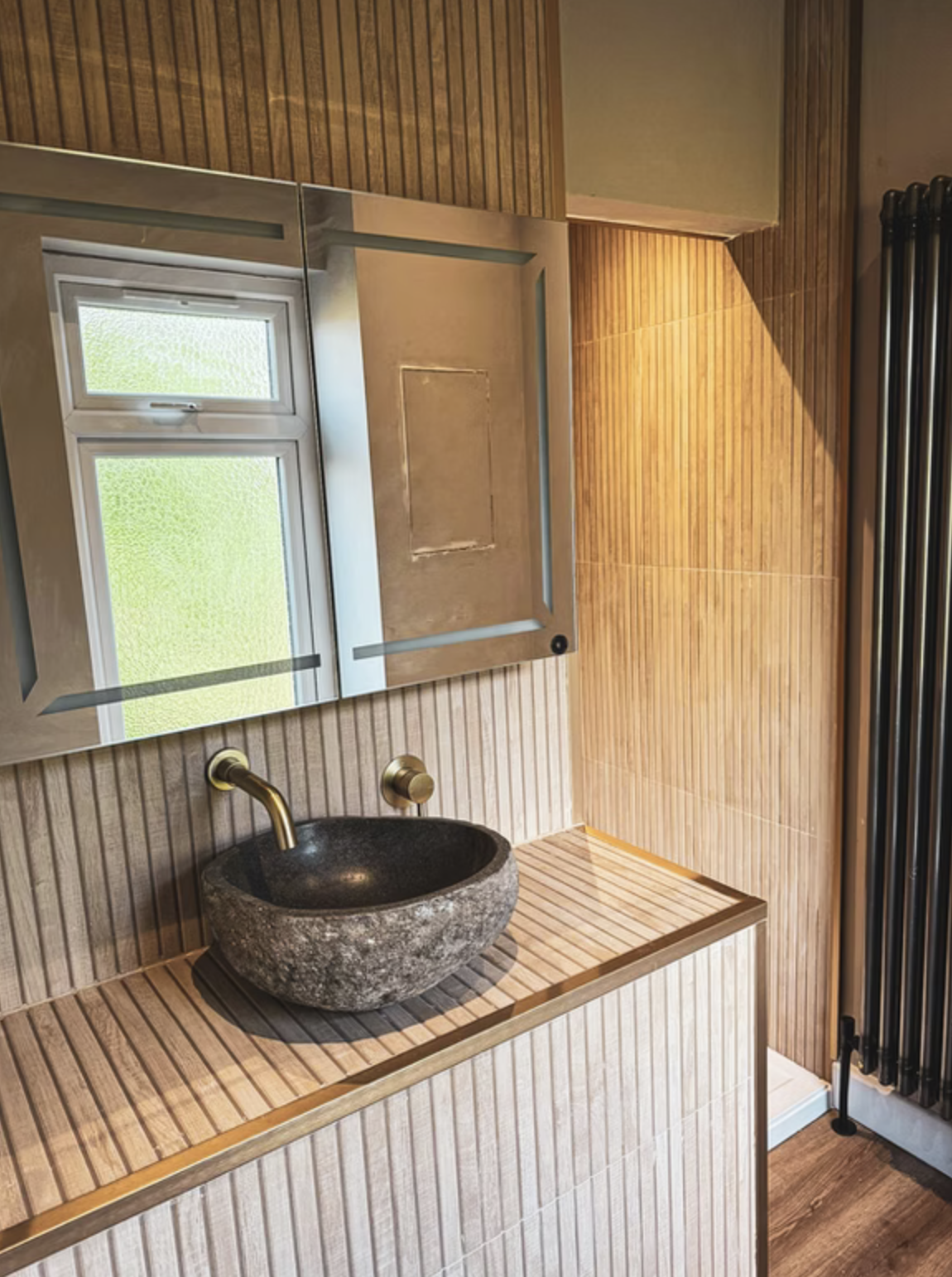 A stone basin with brushed brass wall-mounted tap on a slatted timber vanity, beneath a backlit mirror cabinet.