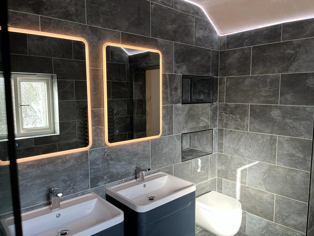 Twin grey vanities, illuminated mirrors, a wall-hung WC and recessed storage within a slate-effect wet room.
